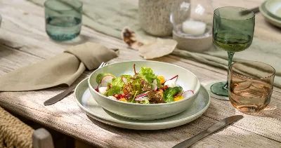 The Perlemor collection from Villeroy & Boch with a fresh salad with various vegetables in a bowl on a wooden table, complete with cutlery, glasses and beige napkins.