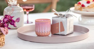 Une bougie parfumée de la collection de like. by Villeroy & Boch sous forme de cadeau sur un plateau rond avec une boîte cadeau argentée en arrière-plan et des orchidées roses au premier plan.