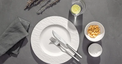 A white plate with fork and knife, a glass of water with lemon, a bowl of crackers and a napkin on a grey table.