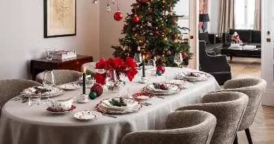 Sala da pranzo decorata per il Natale con un tavolo per otto persone. La tavola è decorata con stoviglie festose, candele e fiori rossi. Accanto è presente un albero di Natale con addobbi.