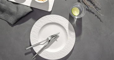 Couverts Piemont sur une assiette Manufacture Assiette blanche vide avec fourchettes et couteaux croisés, un verre d'eau avec du citron vert et une serviette de table sur une surface grise.