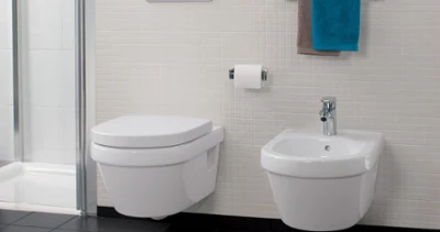 Biała toaleta i bidet Architectura marki Villeroy & Boch Nowoczesna łazienka Architectura z WC montowanym na ścianie, bidetem, uchwytem na papier toaletowy i uchwytem na ręczniki z niebiesko-brązowym ręcznikiem. Podłoga pokryta jest czarnymi płytkami, a ściana małymi kwadratowymi białymi płytkami.