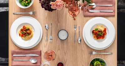 Esstisch mit zwei Royal Tellern Pasta, zwei Salaten, Besteck, einer Kerze und einem Getränk. Esstisch mit zwei Royal Tellern Pasta, zwei Salaten, Besteck, einer Kerze und einem Getränk. Die Villeroy & Boch Teller stehen auf rosa Servietten und auf dem Tisch steht ein rosa Blumenarrangement.