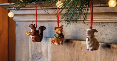 Tres adornos navideños de animales de la colección Nostalgic Ornaments, que incluyen una ardilla, un zorro y un conejo, cuelgan de una guirnalda de pino con cálidas luces de hadas sobre una superficie de madera.