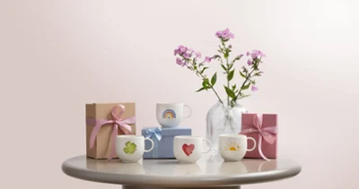 With love mugs with motifs such as hearts, clover leaves, suns, and rainbows Four decorated with love mugs with various motifs such as hearts, clover leaves, suns, and rainbows, two gift boxes, and a vase with pink flowers on a round table in front of a light background.