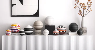 Various La Boule pieces in different sizes and colors A collection of colorful, striped, and patterned spherical La Boule pieces stands on a white console beneath abstract art and a vase of orange flowers.