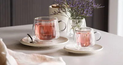 Artesano glass cups from Villeroy & Boch filled with pink tea, placed on saucers next to a spoon on a table that is decorated with a small vase of purple flowers.