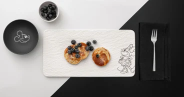 Deux crêpes aux myrtilles sur un plat de service blanc rectangulaire Manufacture Disney Deux crêpes aux myrtilles sur une assiette rectangulaire blanche Manufacture Disney, à côté d'un bol de myrtilles et d'une fourchette posée sur une serviette.
