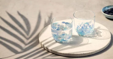 Zwei Like Aqua Wassergläser mit blauen Blütenblättern auf einem runden Tablett, mit Palmblattschatten auf einer hellen Fläche.
