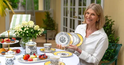 Isabelle von Boch in a white blouse sits at a table outside and sets out a set of decorative Audun plates. The table is set with teacups, flowers, and various baked goods.