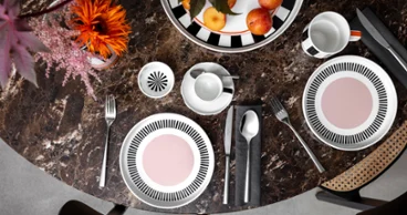 A round marble table with two Memphis place settings, each with a plate, cutlery and a cup. A bowl of apples and a Memphis vase with flowers adorn the center.