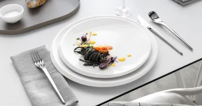 Ein elegantes Tischarrangement mit weißen Tellern und Speisen aus der NewMoon Kollektion, stilvoll arrangiert mit Besteck.

