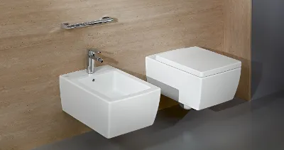 Bagno moderno con WC e bidet bianchi a parete davanti a muri con piastrelle beige, dotato di rubinetteria cromata, sciacquone ed elegante portasciugamani.