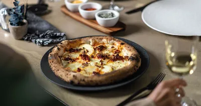 A freshly baked pizza on a black Manufacture plate, accompanied by various condiments and a glass of white wine.