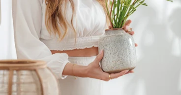 Lave Home Vase as an ideal gift A woman in white clothing hands over a Lave ceramic vase with green and yellow flowers, which stands on a wooden table.