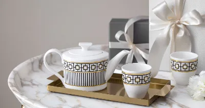 A white and gold patterned teapot and cups from MetroChic on a golden tray on a marble table. Two wrapped presents and a small white flower can be seen in the background.