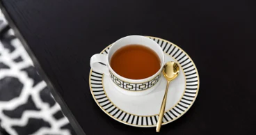 A MetroChic teacup with a golden spoon on a matching saucer, placed, on a dark wooden table.