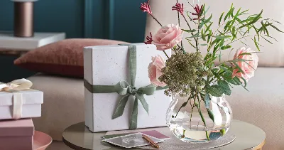 A Rose Garden vase with pink flowers and a gift on a table.