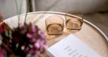 Due bicchieri tumbler minimalisti in cristallo Like Glass con linee arrotondate nella calda tonalità ambra Clay di Villeroy & Boch, posizionati su un tavolino in legno chiaro accanto a un libro bianco e un bouquet sfocato di fiori bordeaux.