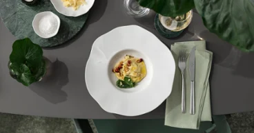 A white Pura pasta plate with pasta and side dishes on a grey table, next to it cutlery, a green napkin and a small bowl of cheese.