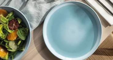 An empty blue Perlemor Aqua plate stands next to a bowl of salad. An empty blue Perlemor Aqua plate stands next to a bowl of salad, cutlery and a napkin on a wooden table.