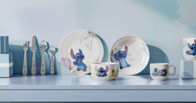 Una serie de platos y tazas de la colección Disney Stitch están colocados en una estantería azul claro. Una serie de platos y tazas de la colección Disney Stitch con motivos de dibujos animados, expuestos en una estantería azul claro junto con otros utensilios.