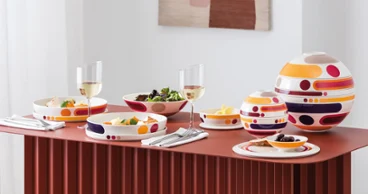 A modern dining table set with colorful patterned dishes, bowls of food, wine glasses, and abstract wall art in the background.