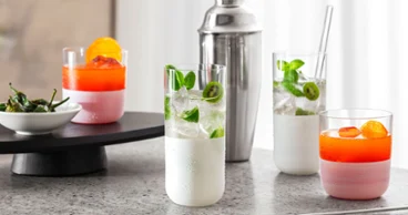 Des verres blancs Manufacture avec des cocktails Quatre cocktails colorés dans des verres Manufacture blancs sur une surface en marbre, avec un shaker à cocktail en arrière-plan et un petit bol de paprika sur un plateau.