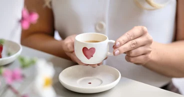 Een persoon houdt een wit koffie kopje met een rood hartmotief boven een bijpassende schotel met rood hartmotief op een tafel.
