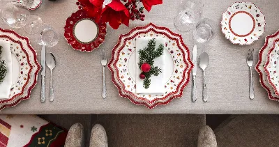 Festive table setting with decorative Toy's Delight plates with red and white patterns, silver cutlery and a centrepiece of red flowers and green plants on a beige tablecloth.