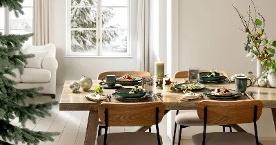A wooden dining table for four with Lave Vert plates, cups, and candles in a bright room overlooking the snow outside.
