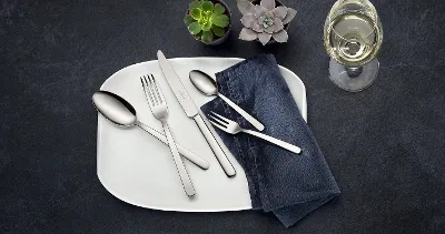 Table setting with a white plate, stainless steel cutlery from the Louis collection, a dark napkin, two small succulents and a glass of white wine on a dark surface.