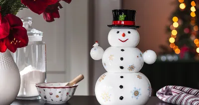 A Santa Claus from Villeroy & Boch with a black hat and red bow on a table next to a sugar bowl, a mixing bowl, and red flowers. Christmas lights are blurred in the background.