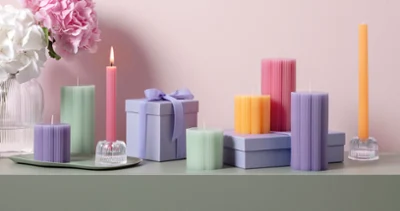 Fleur Dekokerzen in verschiedenen Formen und Farben Eine Sammlung bunter Dekokerzen, der Kollektion Fleur, in verschiedenen Formen und Pastelltönen steht auf einer grünen Fläche, mit rosa Blumen im Hintergrund vor einer hellrosa Wand.