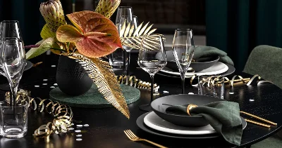 Elegante tavola imbandita di Villeroy & Boch con piatti scuri Manufacture, posate dorate, tovaglioli verdi e un vaso nero Collier con fiori e foglie tropicali. Festoni dorati e bicchieri decorano un tavolo nero.