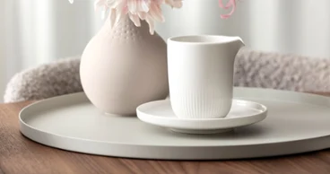 A white ceramic pitcher on a saucer stands on a round tray next to a vase with pink flowers on a wooden table. 