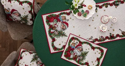 A round table with a green tablecloth is decorated with Christmas placemats and cushions from the Toy’s Fantasy collection with Santa Claus motifs. There are also candles and a decorative white plate on the table.