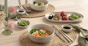 A table set for two with Perlemor pieces A table set for two with bowls of ramen, sushi rolls, edamame, chopsticks and dipping sauces from the Perlemor collection on woven placemats.