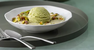 A Pura dessert bowl with pistachio ice cream A Pura dessert bowl with pistachio ice cream, chopped pistachios and cream on a white plate with cutlery beside it.