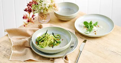 The Perlemor Alga dinner set for one, featuring a plate filled with fresh zucchini pasta, rests on a brown linen tablecloth atop a rustic wooden table, accompanied with silver cutlery.