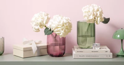 Zwei Fleur Glasvasen mit weißen Hortensien auf einem Tisch Zwei Fleur Glasvasen mit weißen Hortensien auf einem Tisch neben gestapelten Büchern, einer Geschenkbox und einer kleinen grünen Lampe.