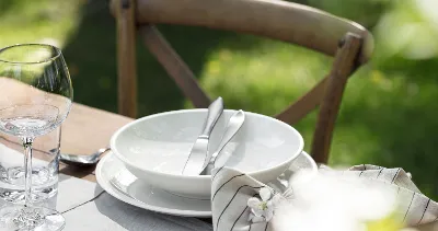 A table setting with a white Artesano plate, a bowl and cutlery, next to a wine glass and a napkin on a wooden table with a chair.