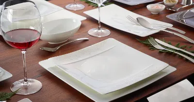 The Modern Grace dining set from Villeroy & Boch consists of square plates, wine glasses, bowls, and flatware arranged on a wooden surface with fern accents. A glass of red wine complements the ambience perfectly.