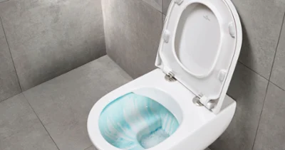 Wisząca miska WC w kolorze białym Toaleta montowana na ścianie z technologią spłukiwania Villeroy & Boch na tle szarych kafelek. Pokrywa toalety jest otwarta i odsłania miskę zawierającą roztwór czyszczący.