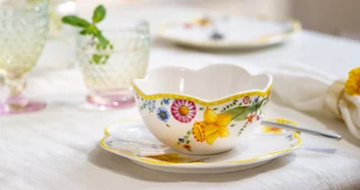 The ‘Spring Awakening’ teacup and saucer with spoon from Villeroy & Boch stand on the table next to crystal glasses and softly blurred flowers in the background.