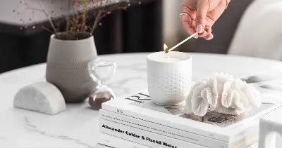 A person lighting a Manufacture scented candle in white on a coffee table with decorative objects and books.