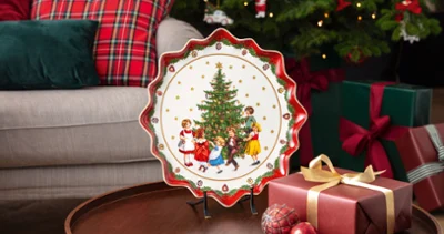 Un plato navideño con niños alrededor de un árbol de Navidad sobre una mesa redonda de madera junto a un regalo envuelto y adornos decorativos. Al fondo, un plato Toy's Fantasy de Villeroy & Boch y cojines a cuadros.