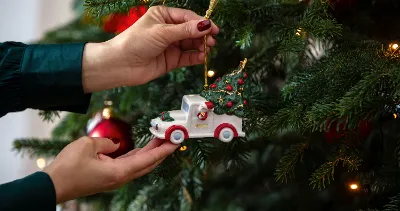 A person hangs a small ornament in the shape of a Villeroy & Boch truck on a decorated Christmas tree adorned with red baubles and lights.