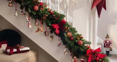 A staircase decorated with green garlands, red bows, and various Christmas decorations from the Nostalgic Ornaments collection such as snowmen and nutcrackers. There are a few wrapped presents on a nearby chair.