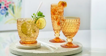 Tre bicchieri in cristallo Boston di Villeroy & Boch nel vivace color arancione zafferano, tra cui un tumbler con lime e menta, un flûte da champagne con alchechengi e un calice da acqua, disposti su un vassoio rotondo in terrazzo su un tavolo bianco.